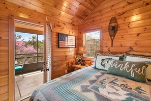 A wooden cabin bedroom with a bed, a window, a door, and a view of pink trees. - Real Log Cabin, Beautiful Screened in Porch, Fireplace, Free Attraction Tickets (Branson)
