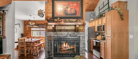 Un interior rústico con chimenea de piedra, zona de comedor de madera y cocina con armarios de madera.