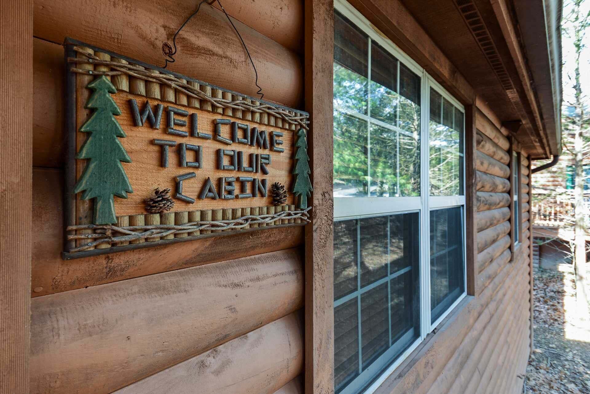 A wooden cabin with a welcome sign featuring a pine tree and pine cones.