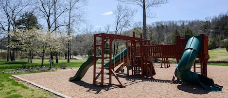 A playground with two slides and a swing set, surrounded by trees and a clear sky.