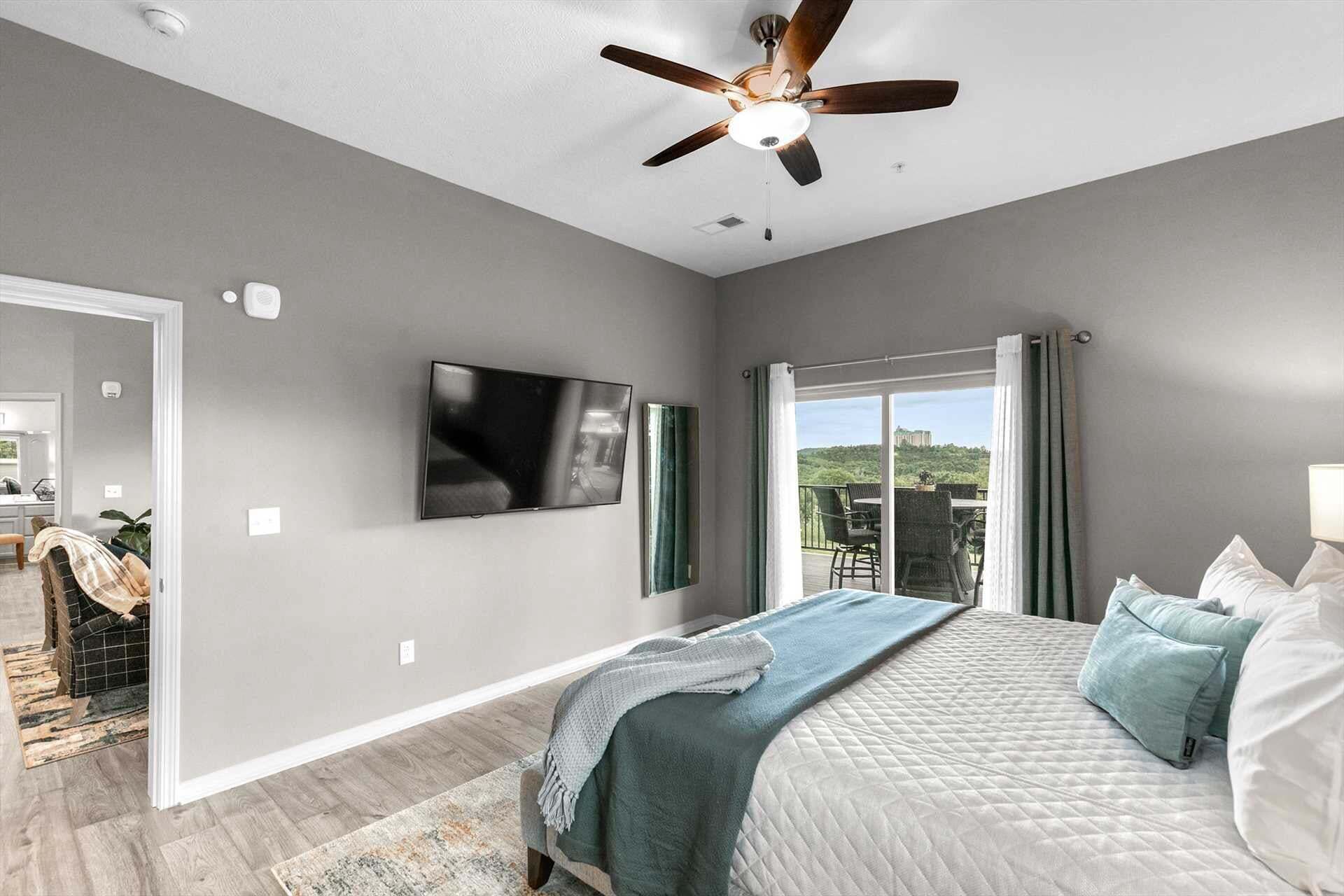 A bedroom with a bed, a ceiling fan, a TV, and a view of the outdoors.