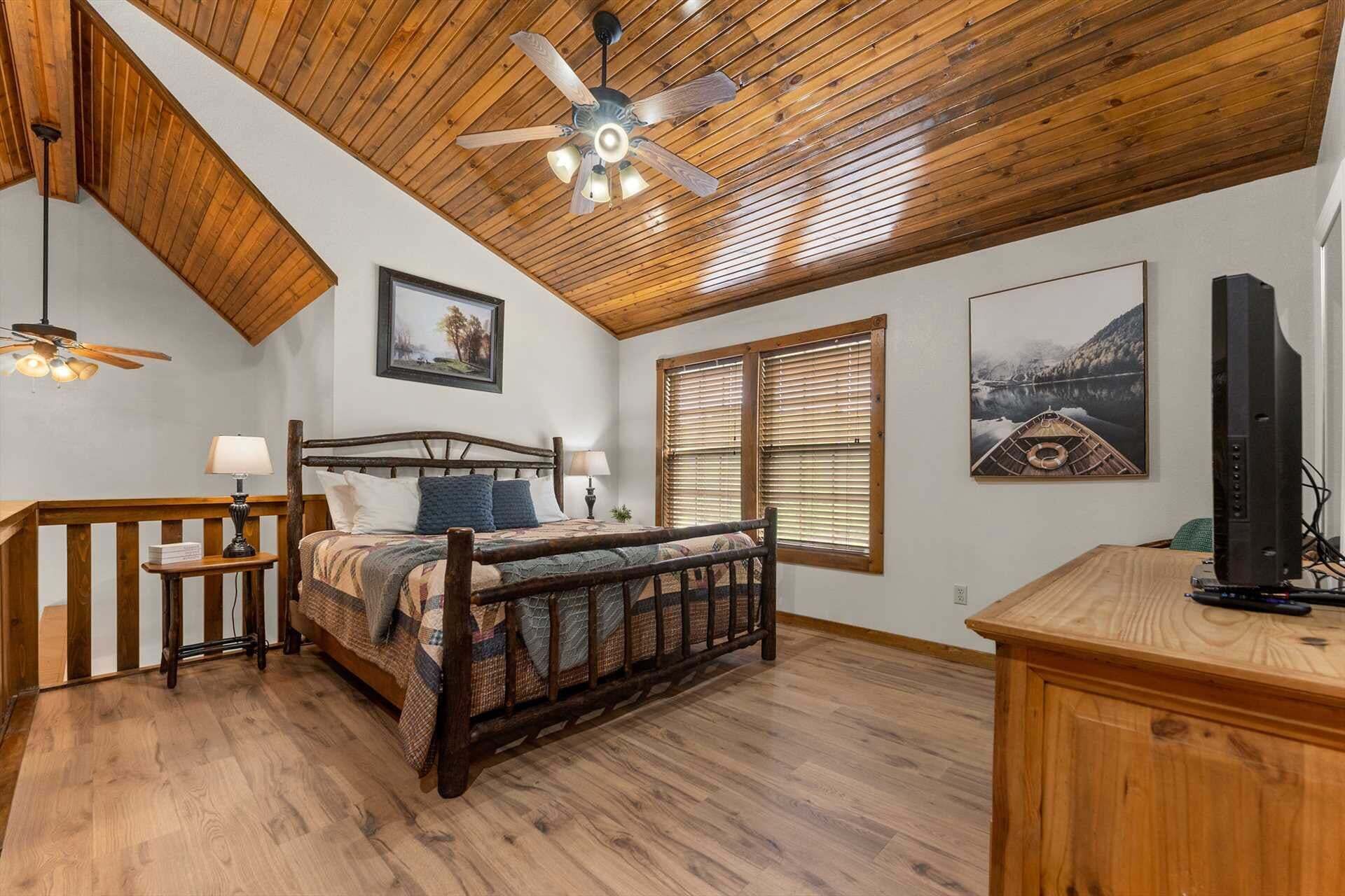 A bedroom with a wooden ceiling, a bed with a quilt, two bedside tables, a ceiling fan, and a television on a wooden stand.