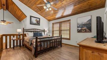 A bedroom with a wooden ceiling, a bed with a quilt, two bedside tables, a ceiling fan, and a television on a wooden stand.