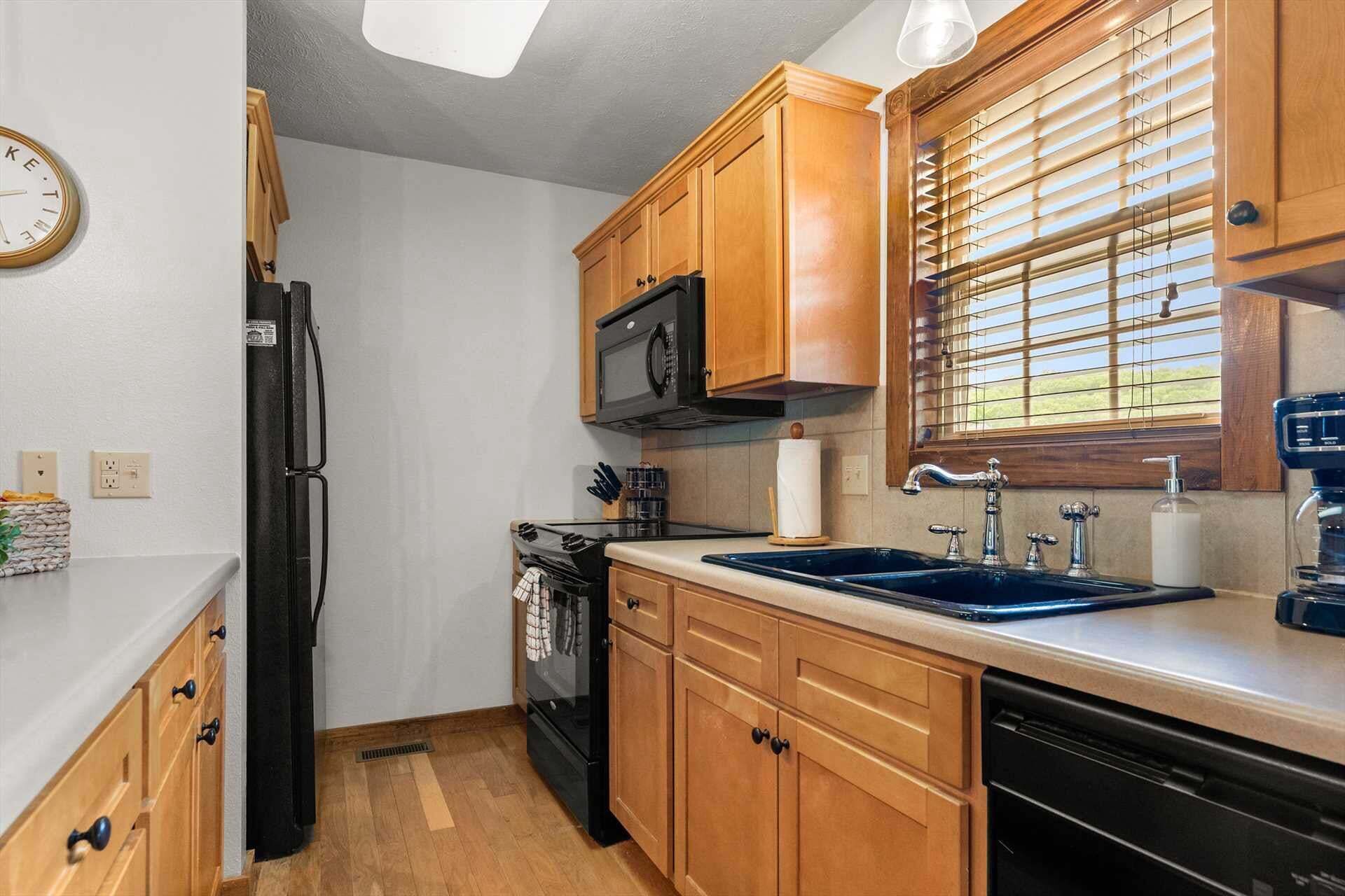 A compact kitchen with wooden cabinets, a black refrigerator, a microwave, a sink with a faucet, and a window with blinds.