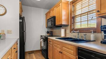A compact kitchen with wooden cabinets, a black refrigerator, a microwave, a sink with a faucet, and a window with blinds.