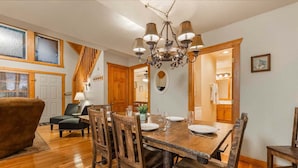 A dining area with a wooden table set for four, surrounded by chairs. A chandelier hangs above the table, and there is a wooden staircase leading to another room. - Private lodge near Silver Dollar City, Beautiful Pool,. Fishing Lake, Ramp Entry, Vaulted Ceilings (Branson West)
