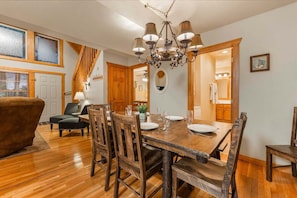 A dining area with a wooden table set for four, surrounded by chairs. A chandelier hangs above the table, and there is a wooden staircase leading to another room. - Private lodge near Silver Dollar City, Beautiful Pool,. Fishing Lake, Ramp Entry, Vaulted Ceilings (Branson West)