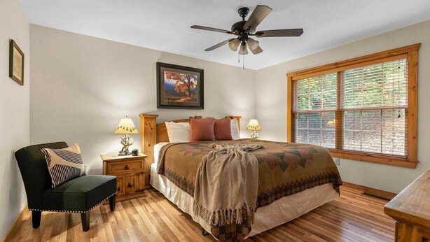A bedroom with a bed, a chair, a nightstand, a lamp, a ceiling fan, and a window. - Private Lodges near Silver Dollar City, Private Lake and Fishing, Beautiful Pool (Branson West)