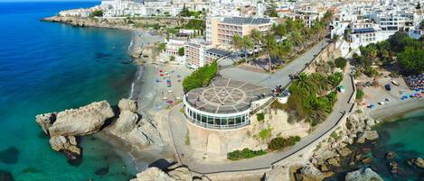 Vista aérea de un pueblo costero con un edificio circular destacado y tejado en forma de estrella, rodeado por un litoral rocoso y aguas azul turquesa.