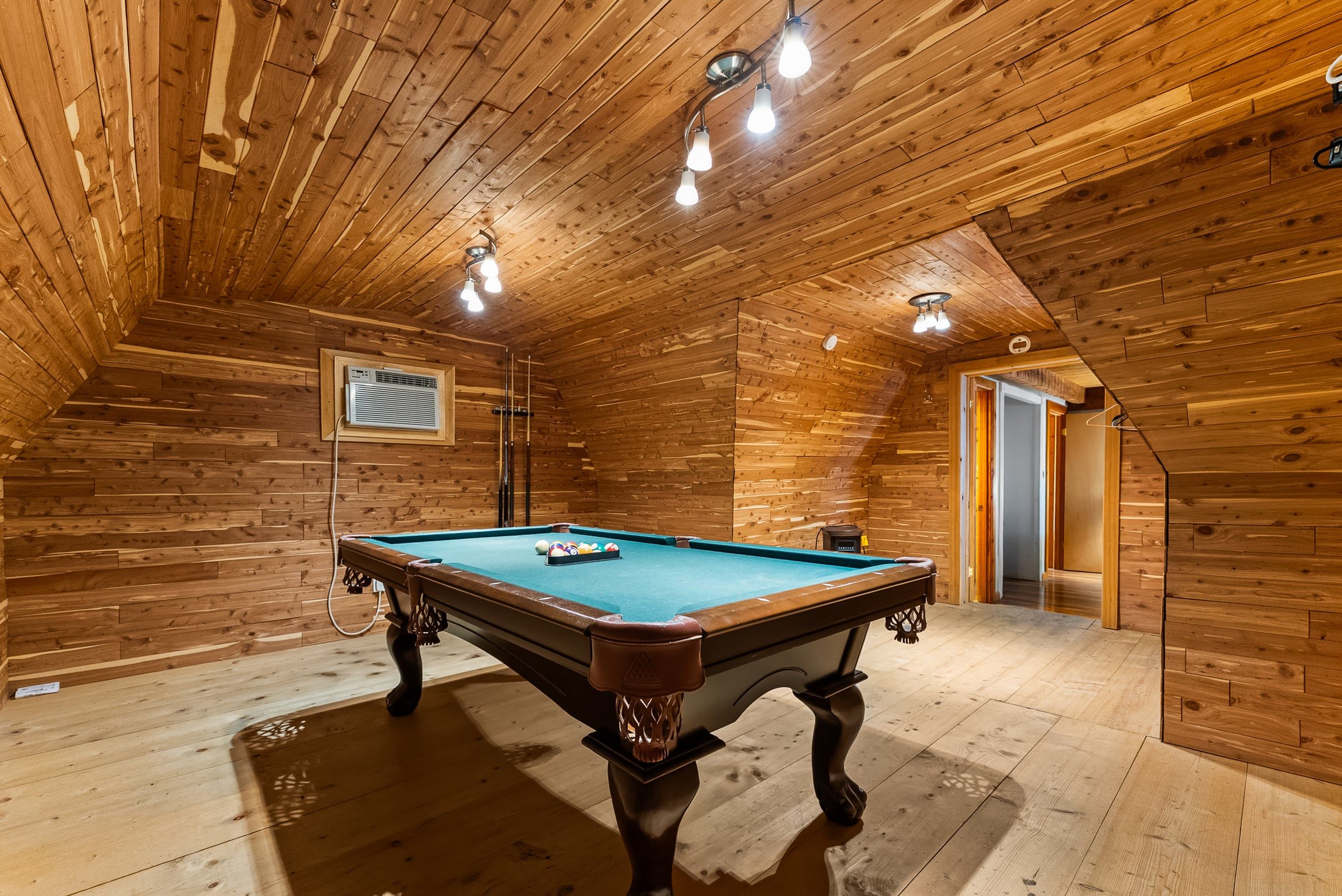 A wooden-paneled room with a pool table, ceiling lights, and a visible air conditioning unit.