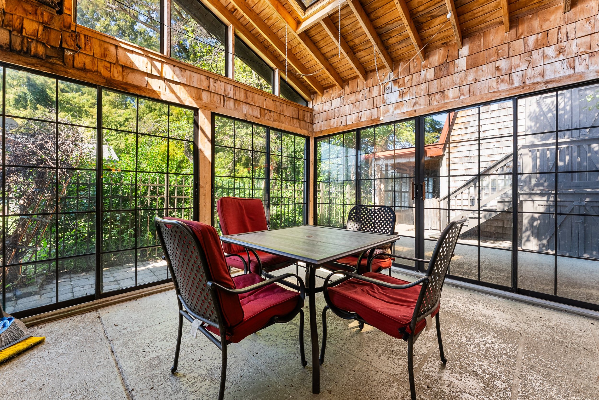 A covered patio with a table and chairs, large windows, and a view of greenery.