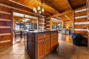 A log cabin interior with a central island, wooden cabinets, a chandelier, and a dining area with a view.