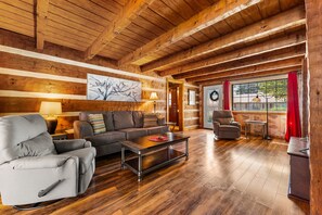 A spacious living room with a wooden ceiling, a sofa, a chair, a coffee table, and a view through a window with a red curtain.
