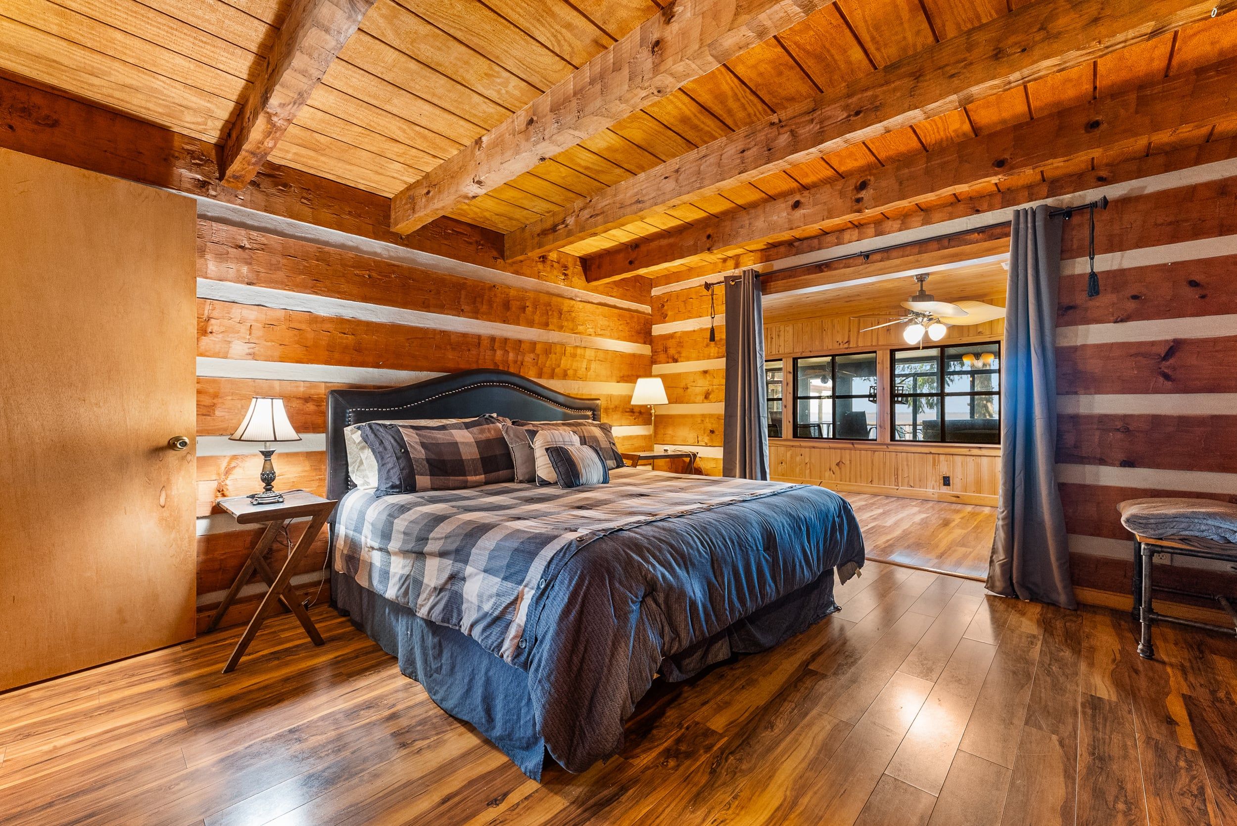 A log cabin bedroom with a bed, a nightstand, a lamp, and a view of the kitchen through a glass door.