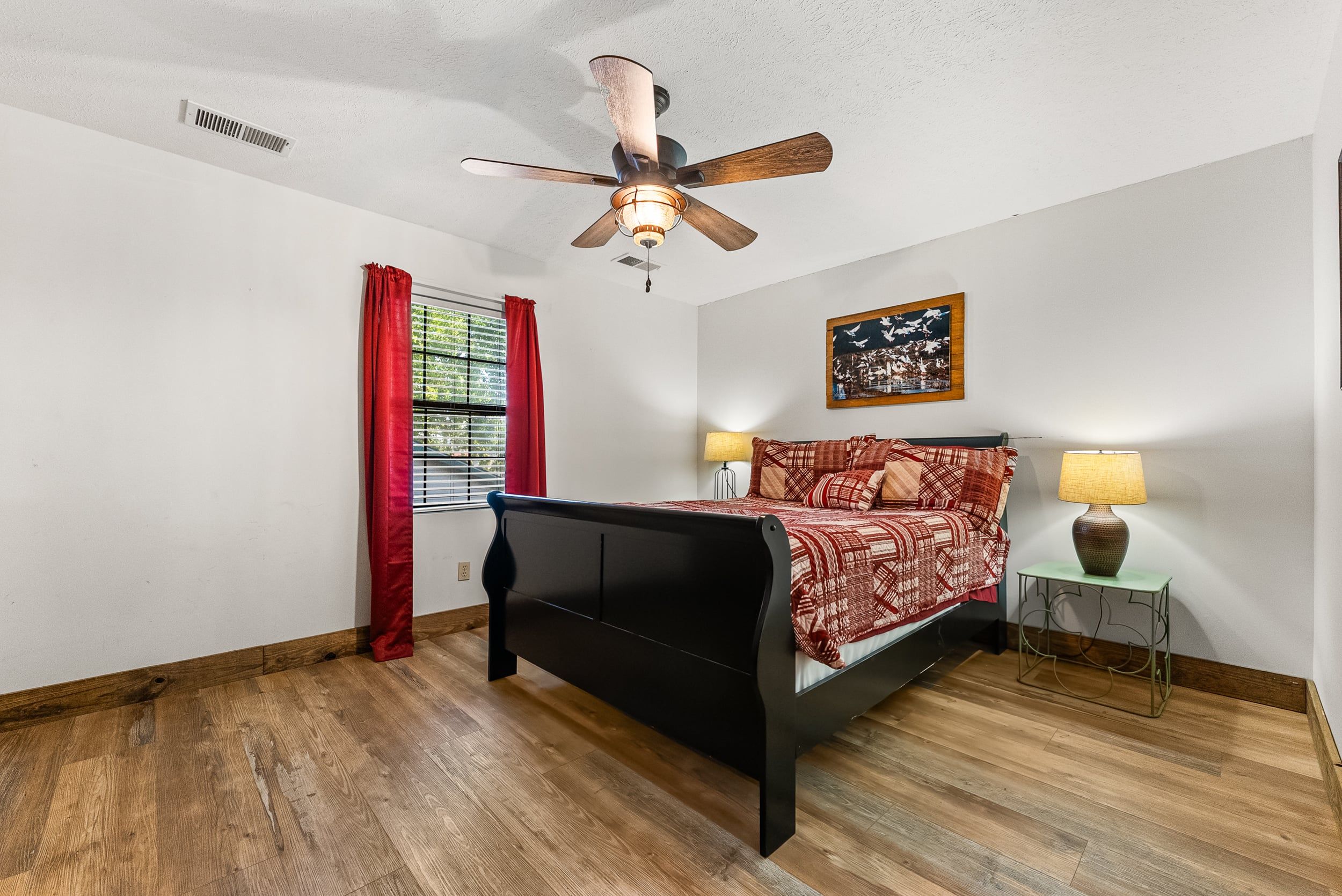 A bedroom with a bed, a ceiling fan, a window with red curtains, a nightstand with a lamp, and a wall-mounted picture.