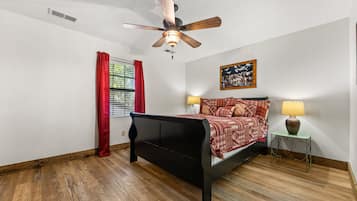 A bedroom with a bed, a ceiling fan, a window with red curtains, a nightstand with a lamp, and a wall-mounted picture.