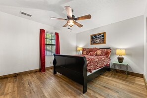 Un dormitorio con cama, ventilador de techo, ventana con cortinas rojas, mesita de noche con lámpara y un cuadro colgado en la pared.