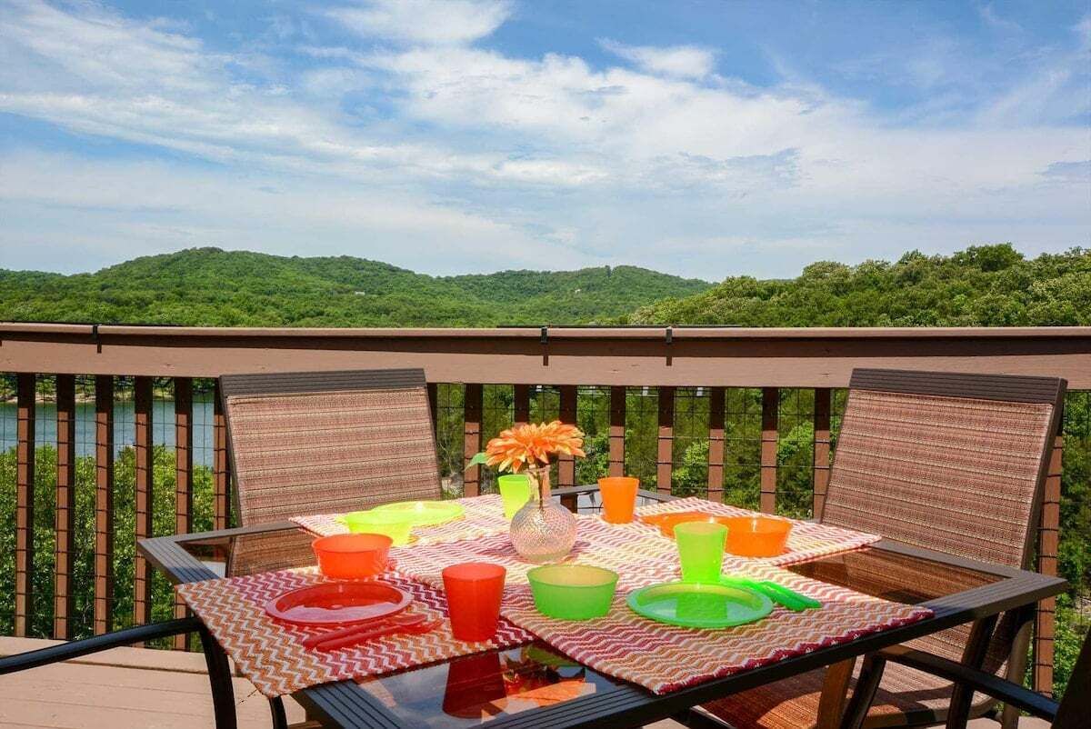 An outdoor dining area with a table set for four, surrounded by chairs, overlooking a scenic view of hills and a body of water.