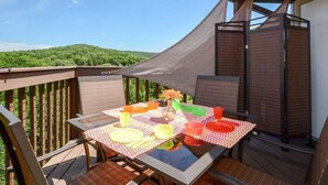 Un área de comedor al aire libre con una mesa dispuesta para dos personas, rodeada por una terraza de madera y vistas a una colina boscosa.