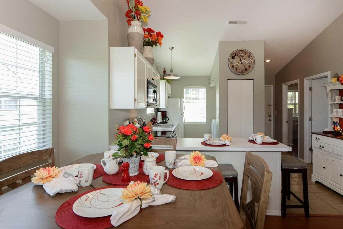 A dining area with a wooden table set for four, a kitchen in the background, and a clock on the wall.