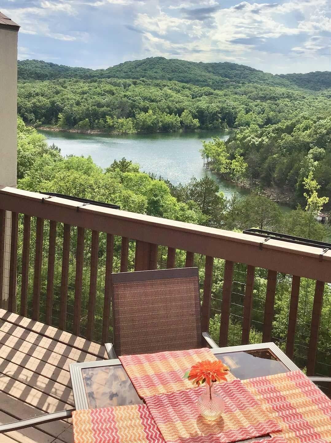 A balcony with a view of a lake surrounded by forest, a chair, and a table with a patterned tablecloth and a flower vase.