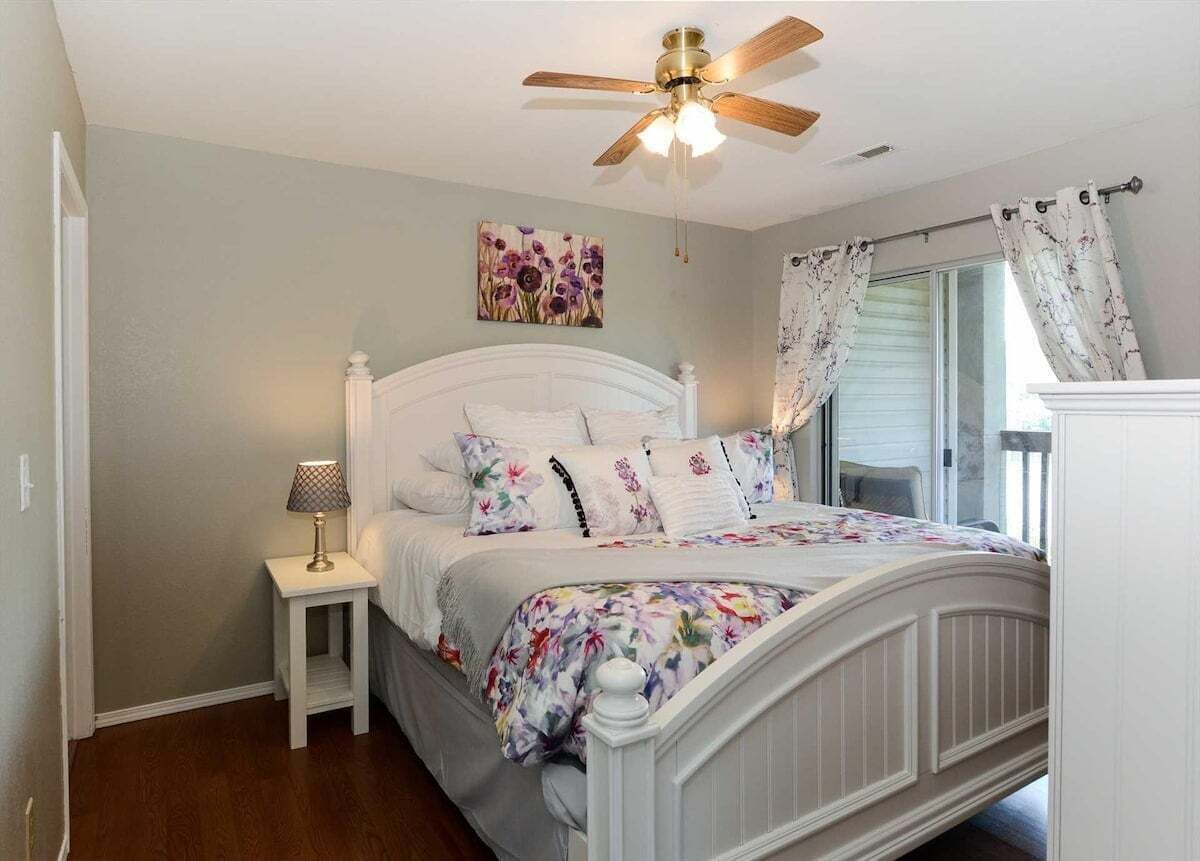 Une chambre à coucher avec un lit, une table de chevet, un ventilateur de plafond et une fenêtre avec des rideaux.