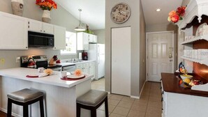 Una cocina moderna con armarios blancos, una isla central con taburetes, un reloj en la pared y una puerta que da a otra habitación.
