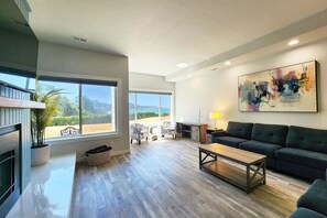 A spacious living area with a large window, a dark sofa, a coffee table, and a painting on the wall. - Ocean Front Condo! Watch for Whales! Walk to Everything! (Depoe Bay)