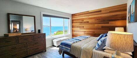 A bedroom with a wooden headboard, a large window with a view of the ocean, a bed with a striped blanket, a dresser, and a bedside lamp.