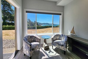 Ein Zimmer mit zwei gemusterten Sesseln, einem kleinen runden Tisch und Blick auf eine Küstenlandschaft durch große Fenster.