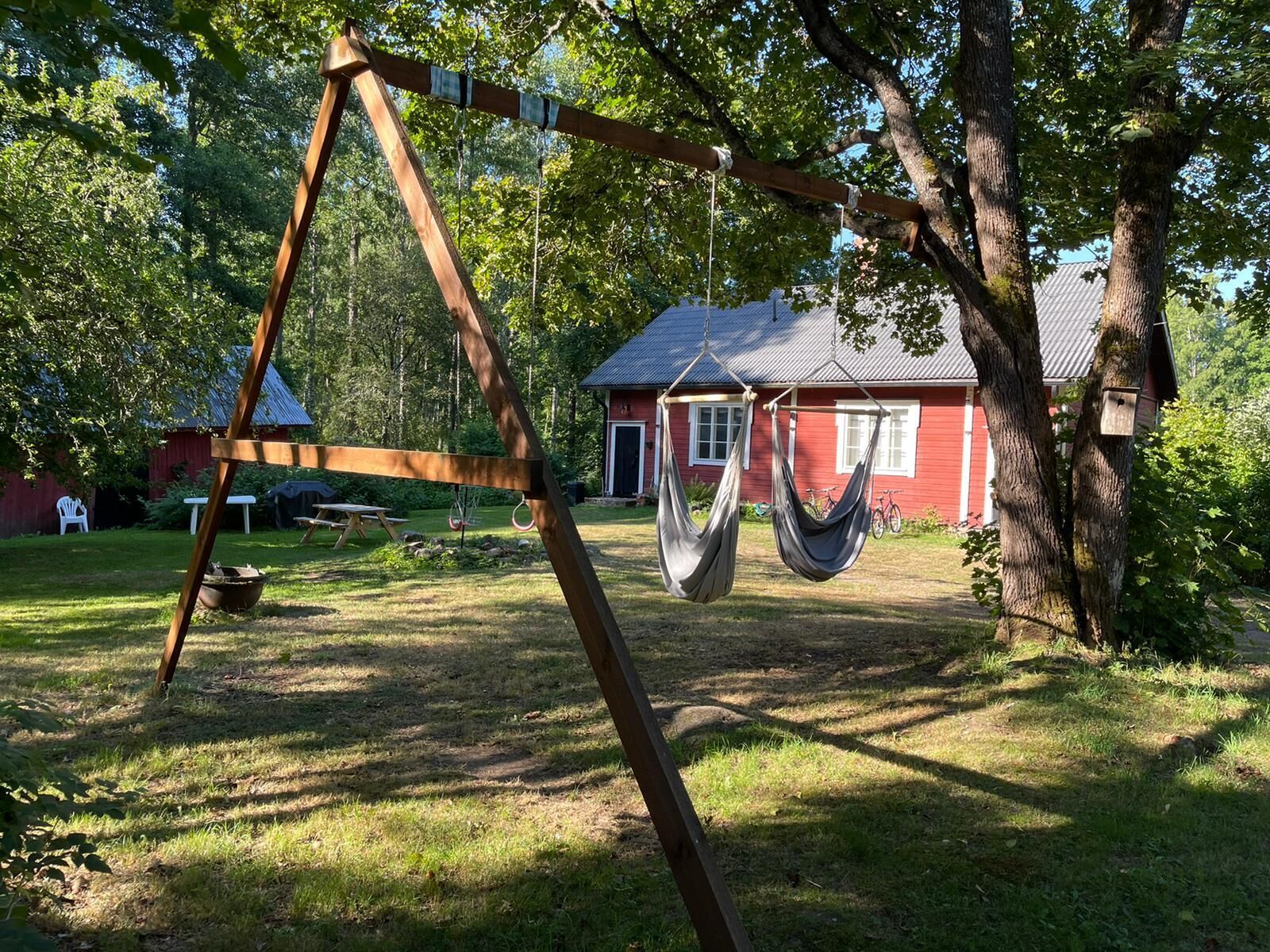 Un hamac suspendu à une structure de balançoire en bois, dans une cour verdoyante, avec une maison rouge en arrière-plan.