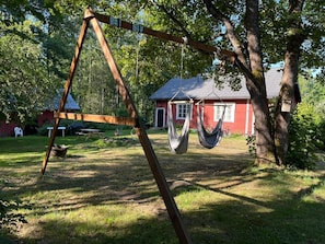 Un hamac suspendu à une structure de balançoire en bois, dans une cour verdoyante, avec une maison rouge en arrière-plan.