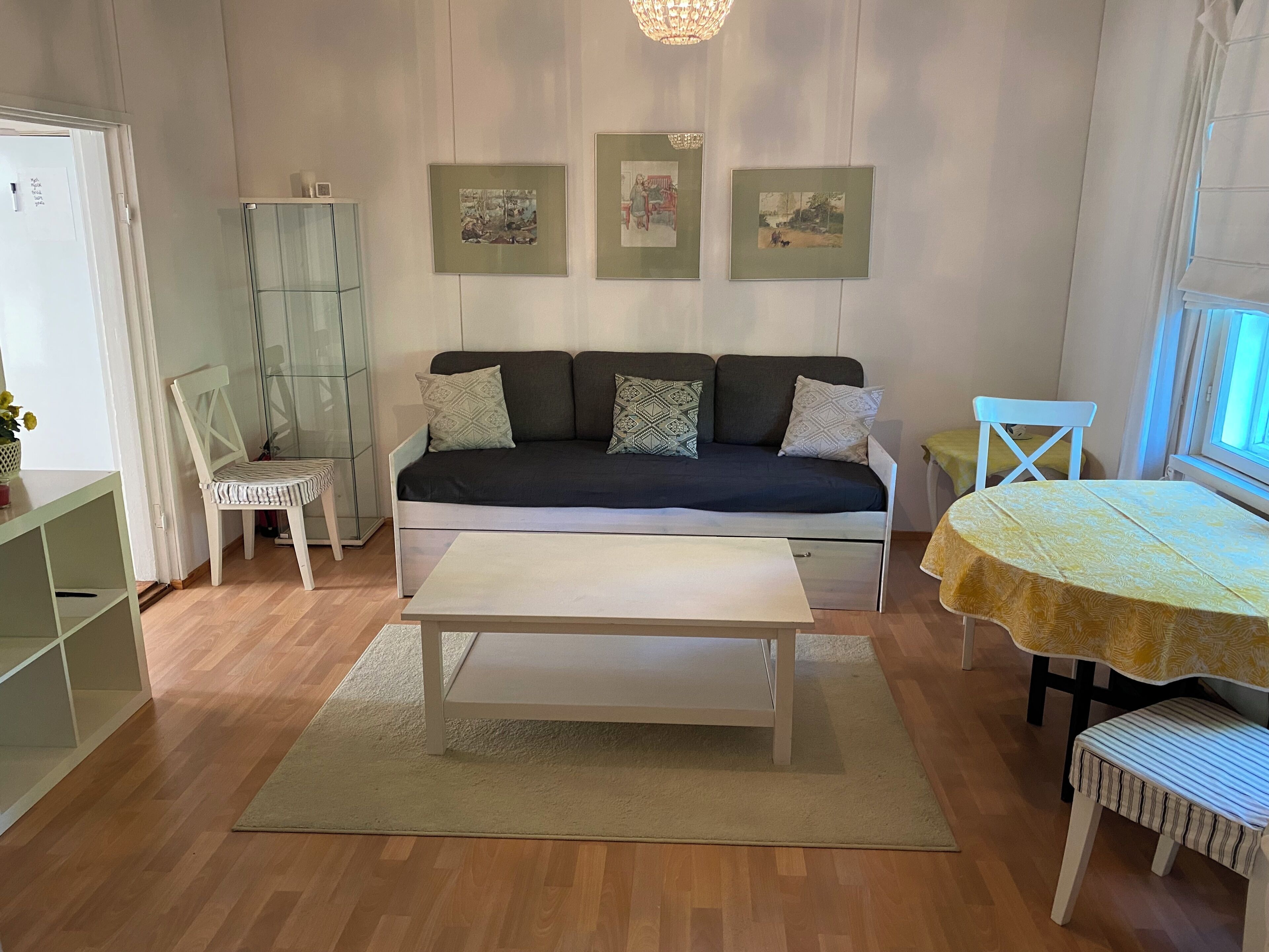 Un salon avec un canapé, une table basse, une table à manger recouverte d’une nappe jaune et une chaise. Des tableaux encadrés ornent le mur, et un lustre est suspendu au plafond.