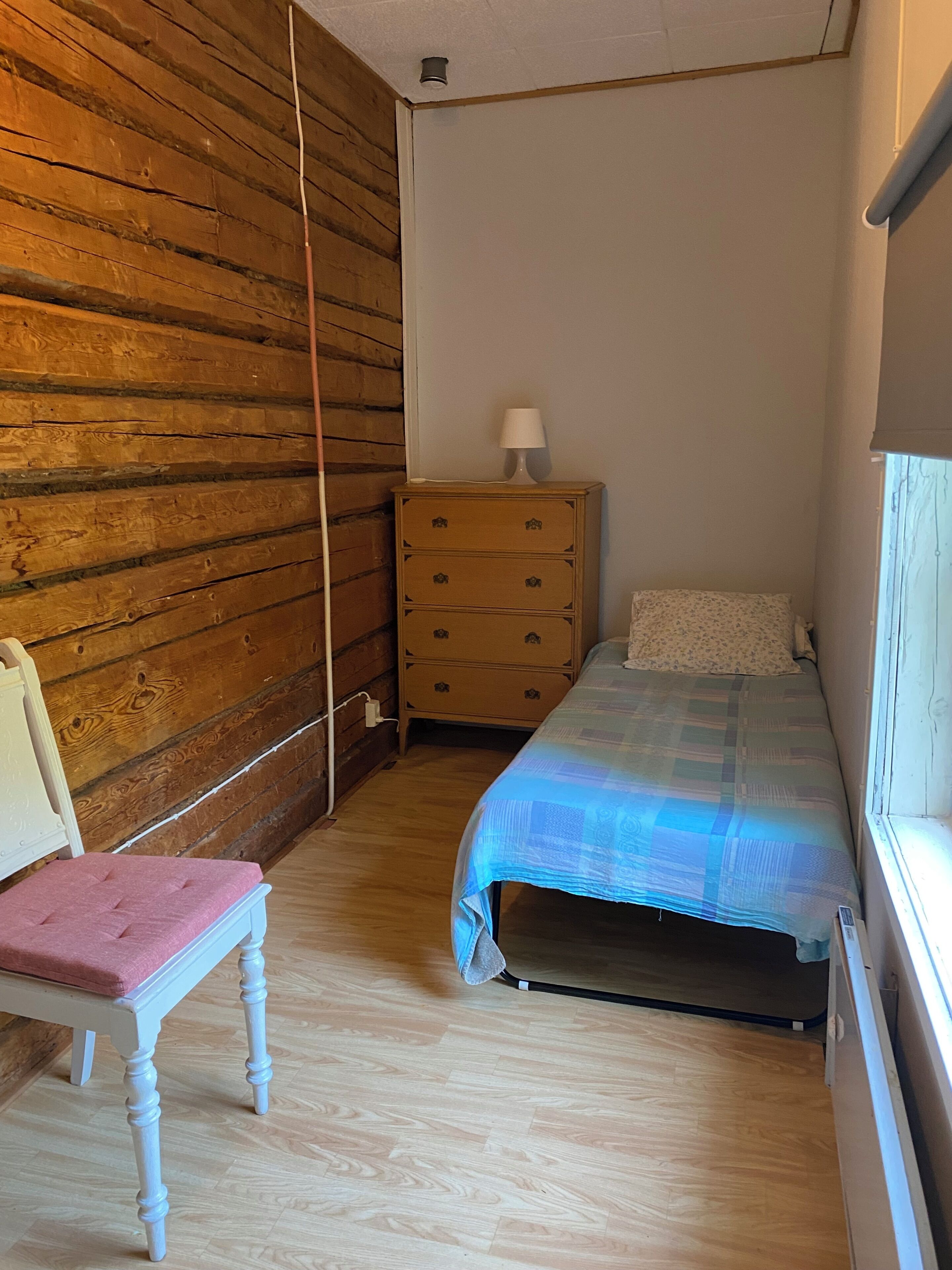 A small bedroom with a wooden wall, a single bed with a blue blanket, a wooden dresser with a lamp, and a chair with a pink cushion.