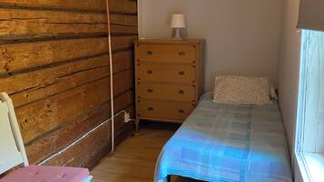 A small bedroom with a wooden wall, a single bed with a blue blanket, a wooden dresser with a lamp, and a chair with a pink cushion.