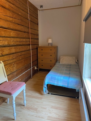 Une petite chambre avec un mur en bois, un lit simple recouvert d’une couverture bleue, une commode en bois avec une lampe et une chaise dotée d’un coussin rose.