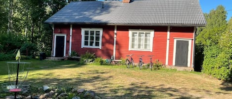 Une maison rouge avec un toit gris, une cheminée et un petit jardin agrémenté d’une clôture en métal et de rochers.
