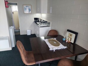 Una pequeña kitchenette con cafetera, un lavamanos y un armario. Hay una mesa con un mantel individual y un marco de fotos.