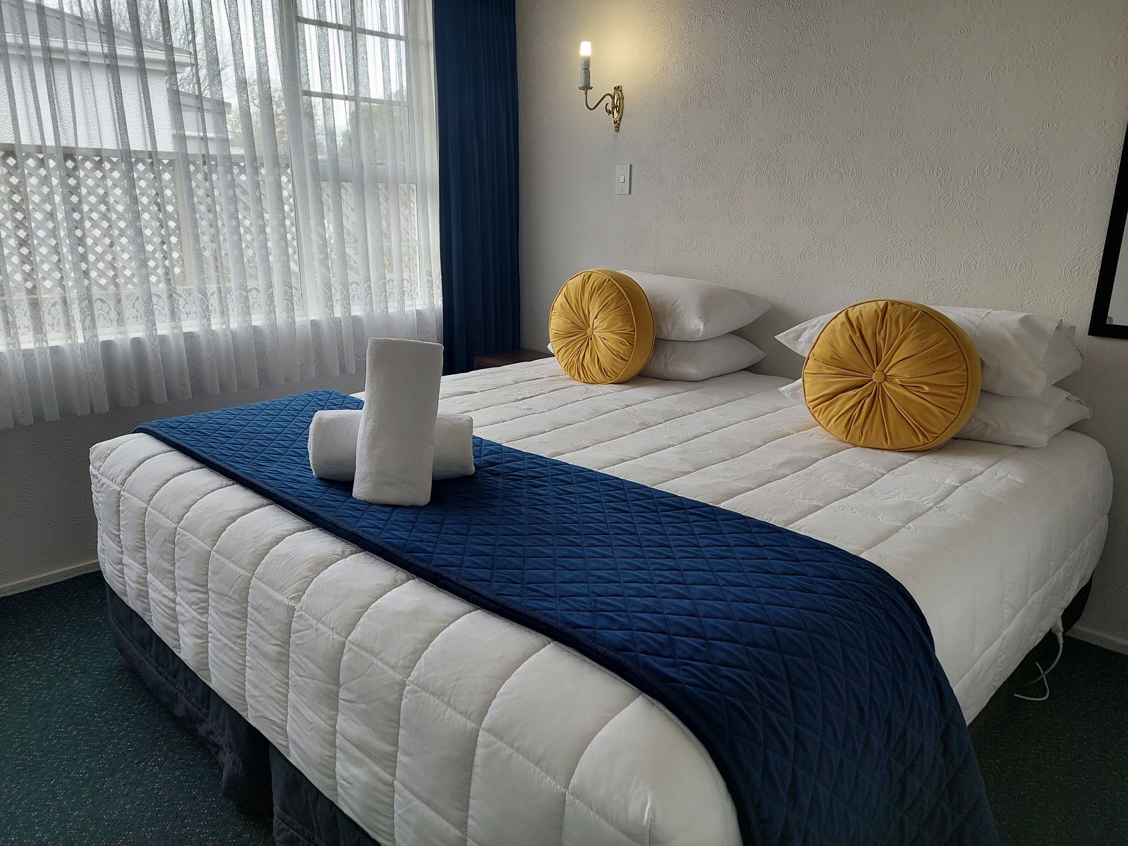 Ein ordentlich bezogenes Bett mit einer blauen Steudecke, weißen Kissen und einem gelben Zierkissen.