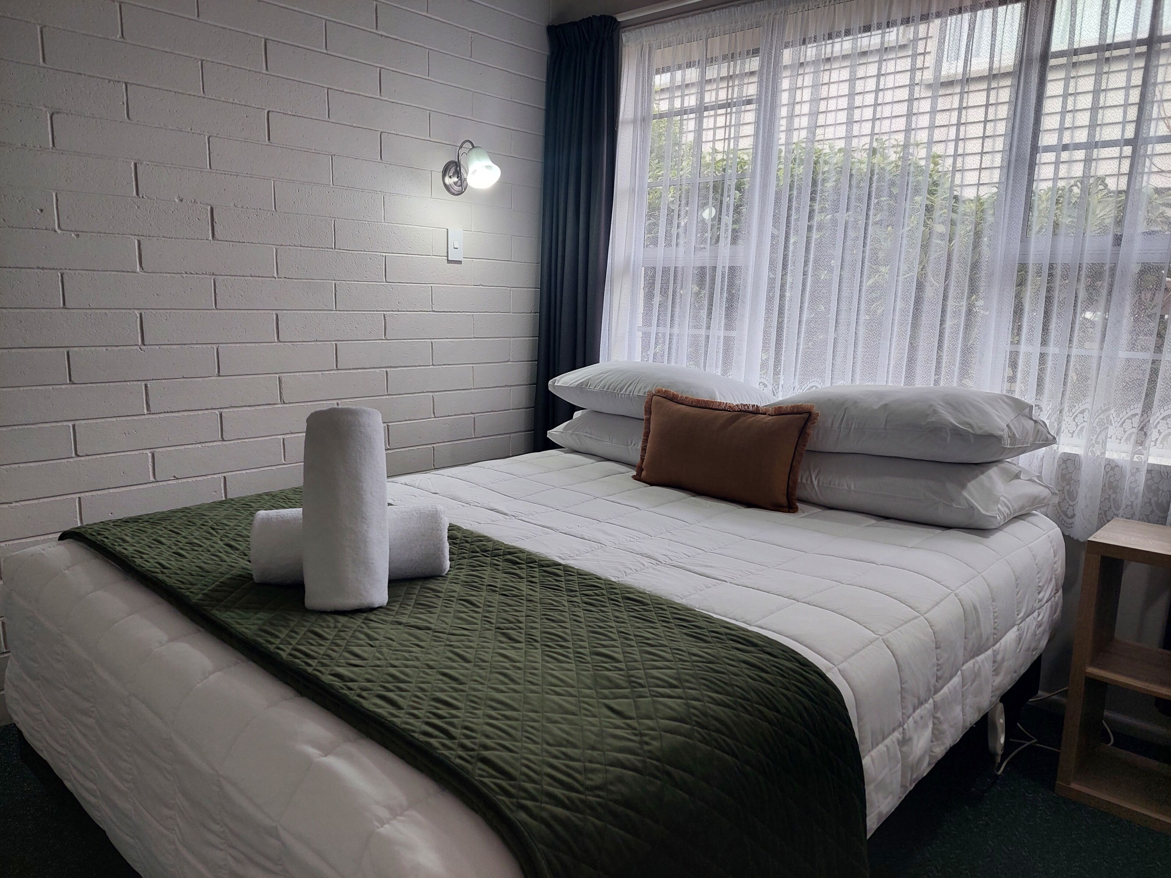 Quarto de hotel com parede de tijolos, uma cama com roupa de cama branca e um edredom verde, um travesseiro marrom e uma janela com cortinas finas.