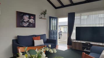 Una sala de estar con un sofá, una mesa con flores, un televisor y una puerta que da al exterior.