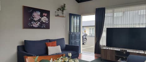 A living room with a sofa, a table with flowers, a TV, and a door leading outside.