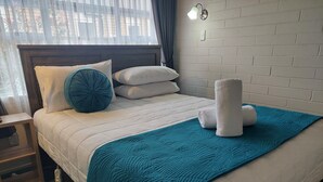 Una cama bien hecha con una manta y almohadillas color turquesa, una mesita de noche con lámpara y una pared de ladrillo.