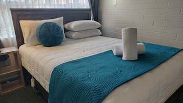 A neatly made bed with a teal blanket and pillows, a bedside table with a lamp, and a brick wall.