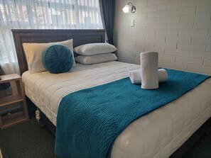 Una cama bien hecha con una manta y almohadillas color turquesa, una mesita de noche con lámpara y una pared de ladrillo.