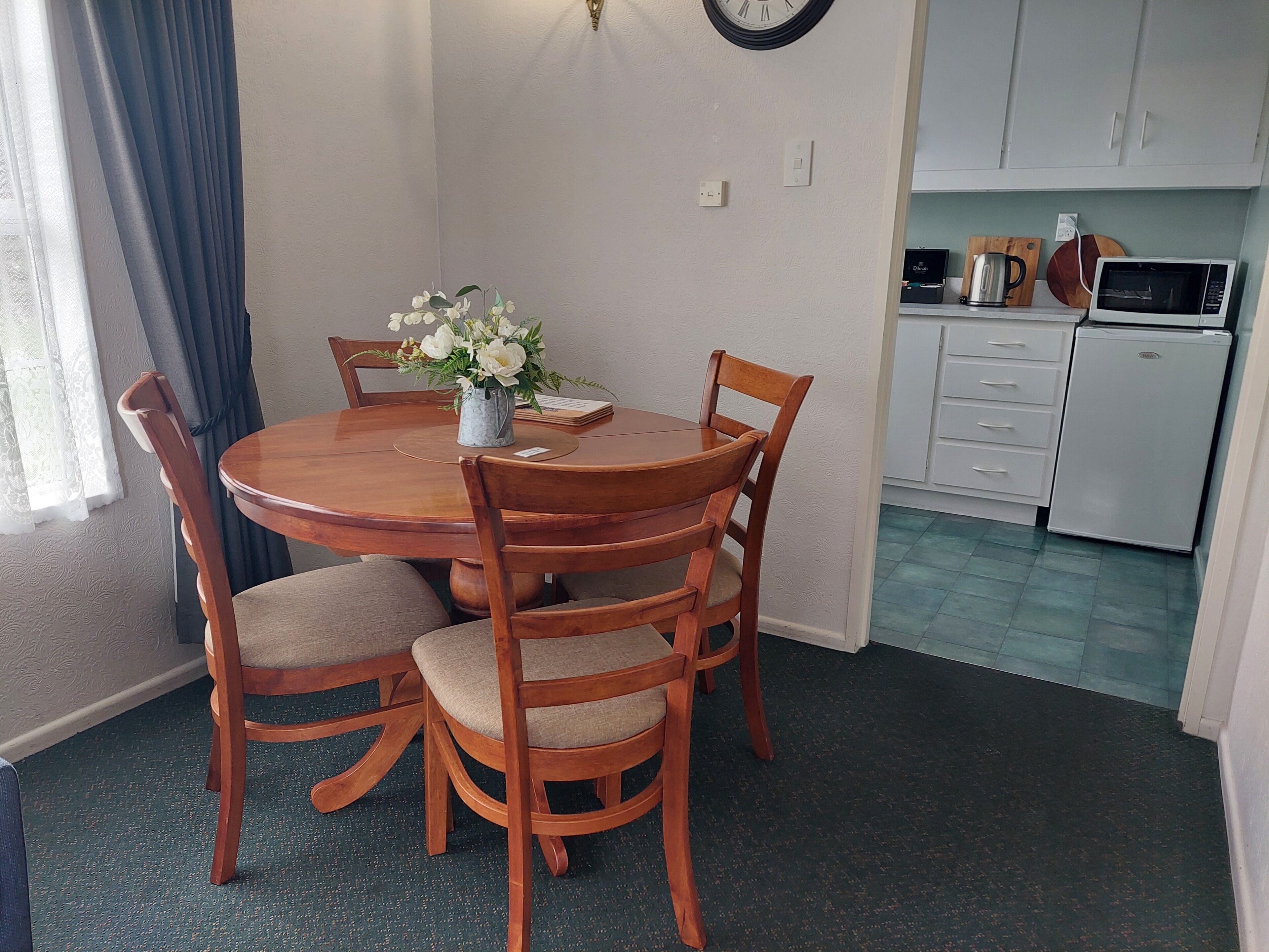 A dining area with a round wooden table, four chairs, a vase with flowers, and a clock on the wall.