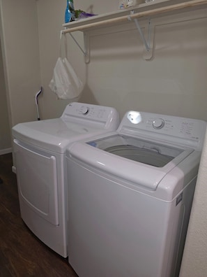 A front-loading white washer and dryer unit with control panels, a white bag hanging on a hook, and a wooden shelf above.