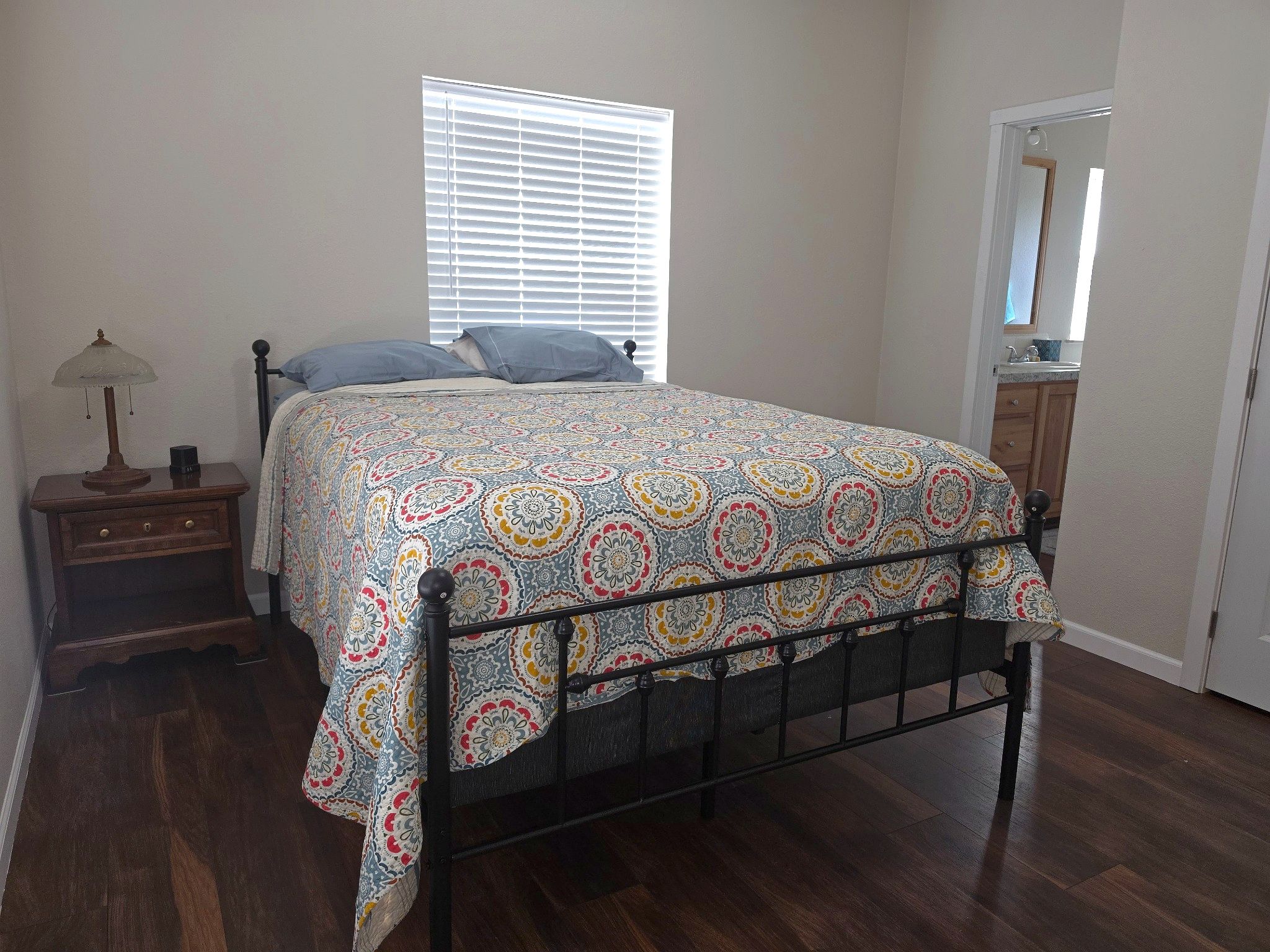 A bedroom with a bed, a nightstand, a lamp, a window with blinds, a door, and a wooden floor.