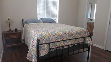 A bedroom with a bed, a nightstand, a lamp, a window with blinds, a door, and a wooden floor.