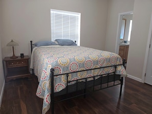 A bedroom with a bed, a nightstand, a lamp, a window with blinds, a door, and a wooden floor.